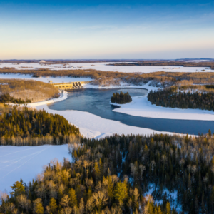Vue aérienne d’un paysage enneigé avec une rivière sinueuse. Un barrage traverse la rivière, entouré de forêts hivernales sous un ciel dégagé symbolisant l'engagement et la réconciliation économique autochtone.