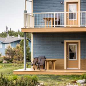 Maison bleue à deux étages avec des portes beige, des balcons et du mobilier d’extérieur. L’herbe verte et les arbres en arrière-plan créent une ambiance paisible.