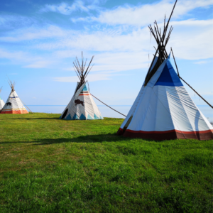 Trois tipis colorés se dressent sur un champ verdoyant sous un ciel bleu clair, avec une eau calme en arrière-plan, créant une atmosphère sereine et paisible.