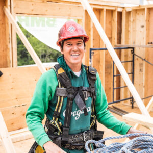 Ouvrier du bâtiment souriant, portant un casque de sécurité rouge et un chandail à capuchon vert, debout sur le chantier. L’ossature en bois et l’équipement de sécurité dégagent une ambiance positive et laborieuse.