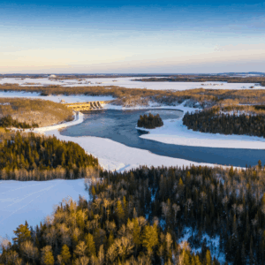 Vue aérienne d’un paysage enneigé avec une rivière sinueuse. Un barrage traverse la rivière, entouré de forêts hivernales sous un ciel dégagé symbolisant l'engagement et la réconciliation économique autochtone.