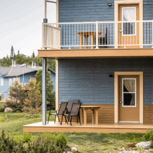 Maison bleue à deux étages avec des portes beige, des balcons et du mobilier d’extérieur. L’herbe verte et les arbres en arrière-plan créent une ambiance paisible.