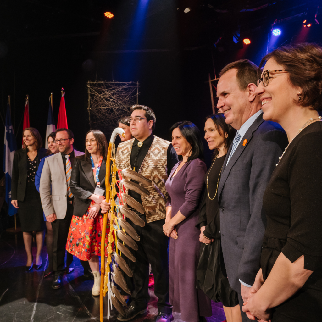 Groupe diversifié de neuf personnes souriantes sur scène, avec drapeaux en arrière-plan, symbolisant l'engagement et la réconciliation économique autochtone.