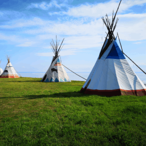 Trois tipis colorés se dressent sur un champ verdoyant sous un ciel bleu clair, avec une eau calme en arrière-plan, créant une atmosphère sereine et paisible.